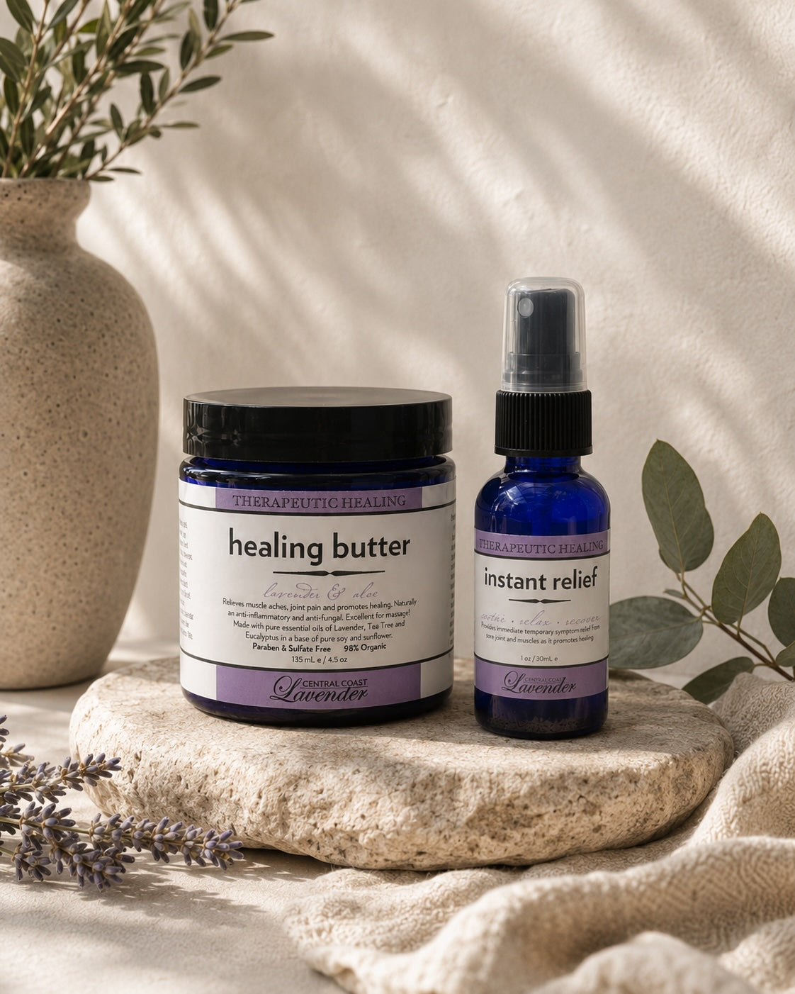 Central Coast Lavender Healing Butter jar and Instant Relief spray bottle side by side on a travertine slab with lavender and eucalyptus in soft daylight. Both labels carry a THERAPEUTIC HEALING band.