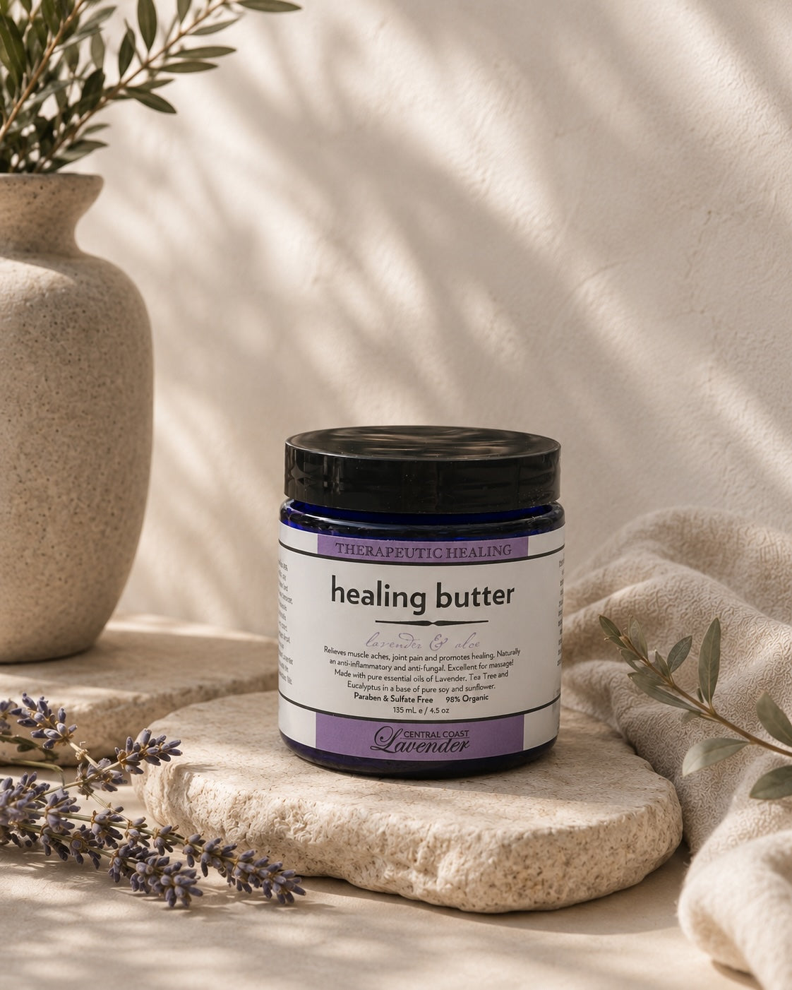 Central Coast Lavender Healing Butter jar on a travertine slab in soft daylight, with lavender sprigs and folded linen. The label band reads THERAPEUTIC HEALING above the words healing butter, lavender and aloe.