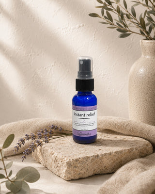 Central Coast Lavender Instant Relief spray bottle on a travertine slab with lavender sprigs, eucalyptus and an olive branch in a stone vase. The label band reads THERAPEUTIC HEALING above the words instant relief, soothe relax recover, 1 oz / 30mL e.